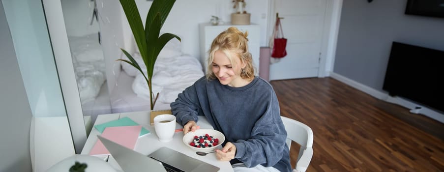 Portrait of smiling blond young woman, eating in front of laptop, watching videos online while having breakfast, enjoying dessert, sitting in bedroom.