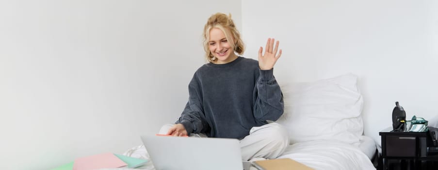 Portrait of young happy woman, connects to video chat, using laptop, waving hand at camera, saying hello to someone online, sitting on bed, studying, e-learning or working from home.