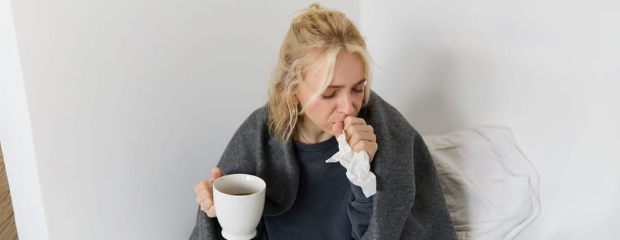 Concept of flu and quarantine. Close up portrait of young woman feeling sick, caught a cold, sneezing and blowing nose in napkin, holding warm tea in hand.