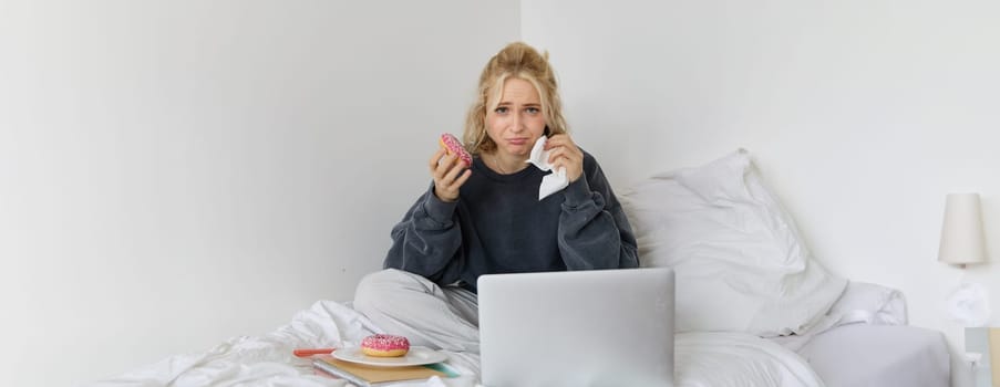 Portrait of sad and depressed woman, feeling heartbroken, lying in bed with comfort food, eating doughnut, crying from upsetting movie, watching something on laptop.