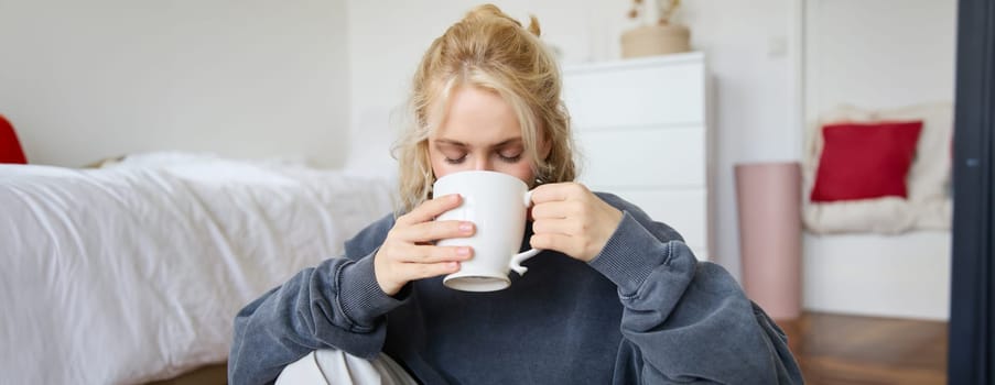 Portrait of young woman sitting on bedroom floor, drinking tea, holding white mug and smiling at camera.