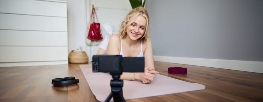 Portrait of young woman, sports vlogger, fitness instructor recording video of herself showing workout exercises, using digital camera, lying on yoga rubber mat.