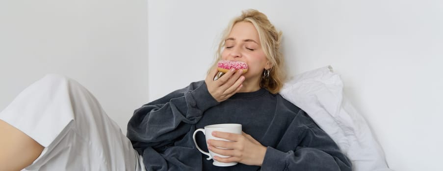 Close up of young blond woman lying in bed with cup of tea, biting delicious doughnut with pleasure and satisfied face.
