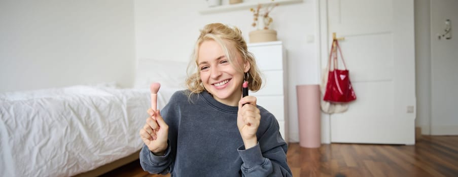 Woman beauty blogger, records video of herself sitting in a room and rating makeup products, puts on make up, holds lipstick and cosmetic brush in hand, using professional camera for content creation.