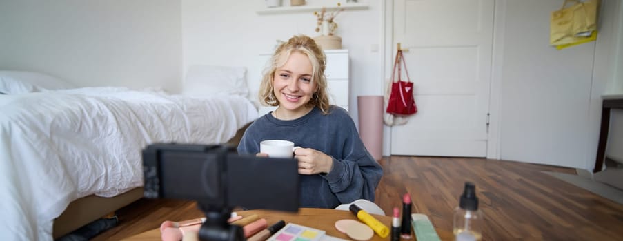 Portrait of young lifestyle blogger, woman sitting in bedroom and talking at digital camera, drinking tea, recording a vlog. Lifestyle content creation