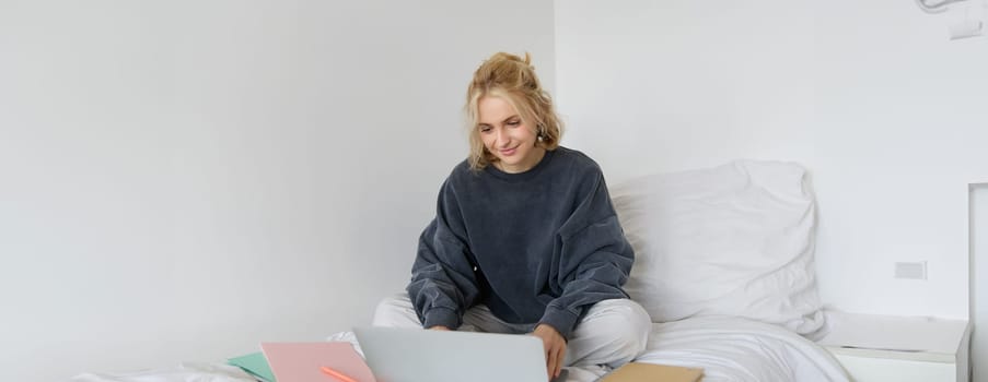 Image of happy young woman, student e-learning from home, connect to online course on her laptop, sits on bed with notebooks, smiling and looking happy.