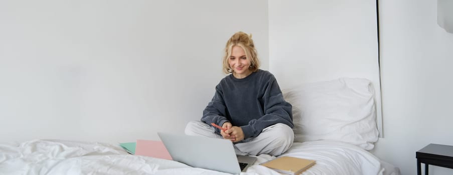 Portrait of happy blond woman, freelancer working from home, sitting on bed with laptop and notebooks. Student doing homework in bedroom, connects to online class via video chat.