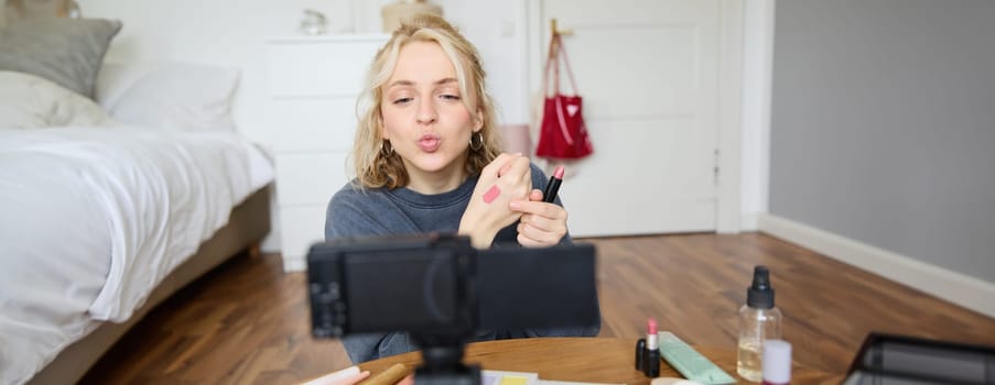 Portrait of young female blogger, beauty makeup vlogger, showing lipstick swatches on her hand, recommending cosmetics to her followers, creating content for social media account on digital camera.