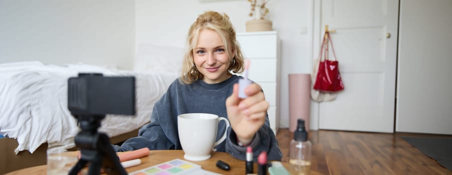 Portrait of smiling charismatic woman, puts on lip gloss, drinks tea, records video on digital camera, beauty content for social media account.