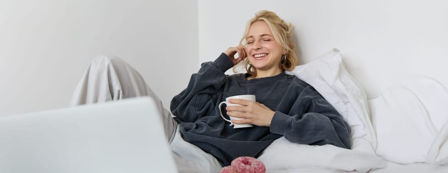 Portrait of happy woman enjoying day-off at home, lying in bed, watching tv show on laptop, looking at screen and smiling, drinking tea and eating in bedroom.