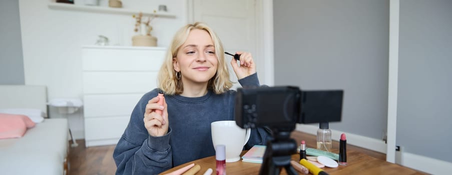 Image of cheerful, beautiful young lifestyle blogger, woman sitting on floor and recording video about makeup, holding mascara, making lifestyle content for her social media account and followers.