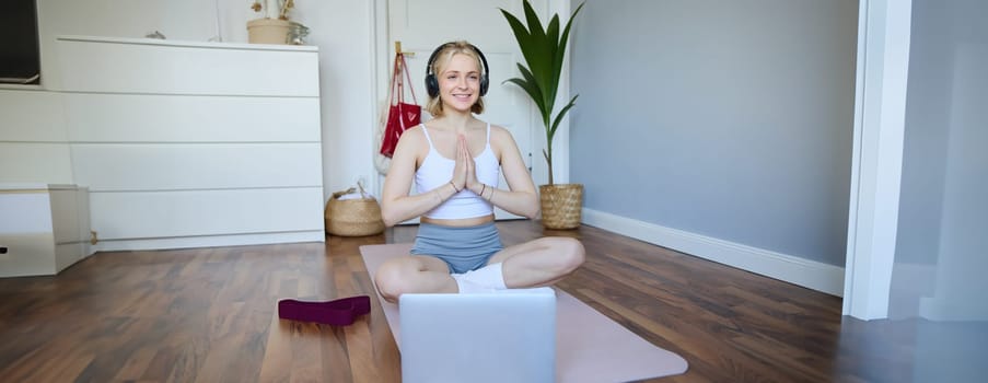 Portrait of blond young woman meditating, practise yoga, using yoga podcast to workout at home, sits on rubber mat.
