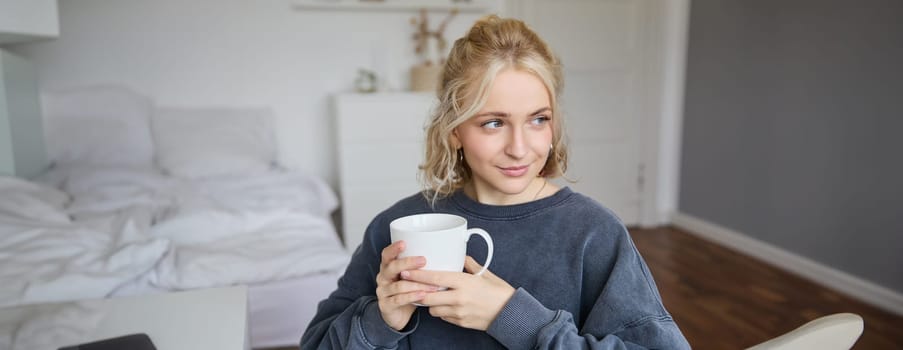 Portrait of smiling beautiful young woman, sitting in bedroom with cup of tea, resting at home alone, enjoys her weekend indoors.