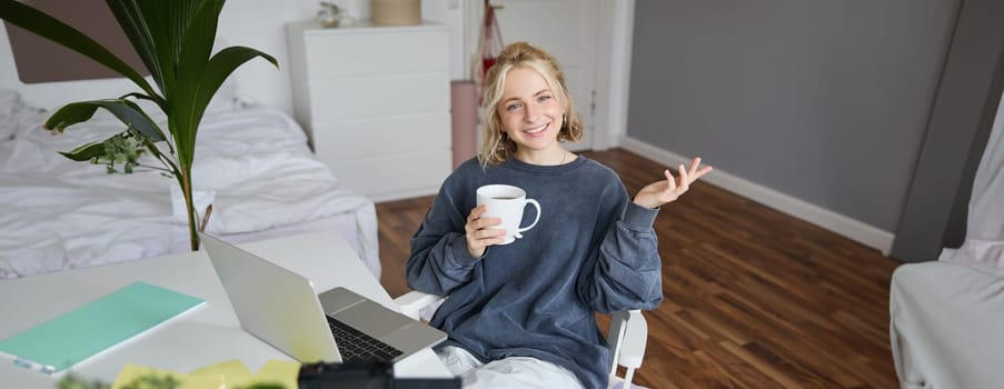 Image of young woman, social media influencer, editing her video on laptop, sits in a room with computer and digital camera, drinking coffee, smiling at camera.