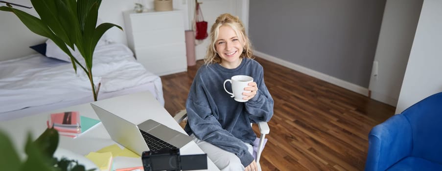 Portrait of young smiling woman, vlogger recording lifestyle video in her room, sits in front of laptop and digital camera, drinks tea, creates content for social media account.