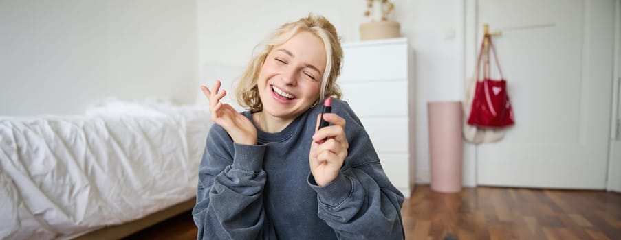 Portrait of smiling beautiful woman in her room, sitting and showing lipstick, recommending favourite beauty product, content maker recording a video of herself for social media blog.