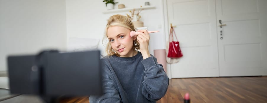 Portrait of young beautiful woman applies makeup blushes with a brush and records herself on digital camera, creates content for beauty blog, social media account.