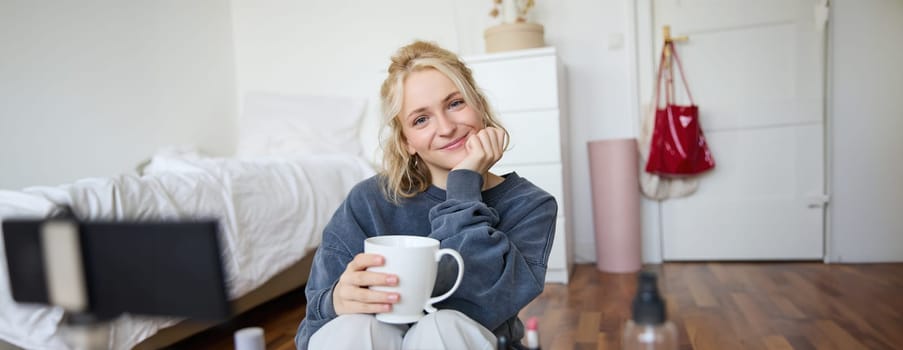 Close up portrait of blond young woman, sitting in her room, looking at camera, holding cup of tea, recording cosy lifestyle blog, vlogger creating content for social media account.