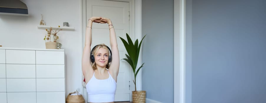 Vertical shot of smiling, active and healthy woman workout at home, follows fitness instructions, watches video on laptop in headphones, stretches her arms.