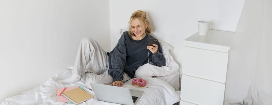Lifestyle and people concept. Young beautiful woman, staying at home, lying in bed with laptop and smartphone, eating doughnut, enjoying free time, spending weekend at home, watching movie online.