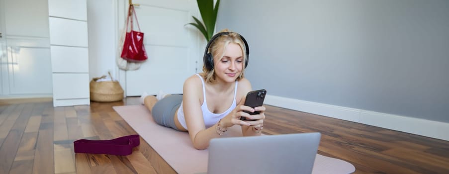Portrait of young woman workout, watching exercise videos on laptop in headphones, lying on rubber mat with mobile phone and smiling.