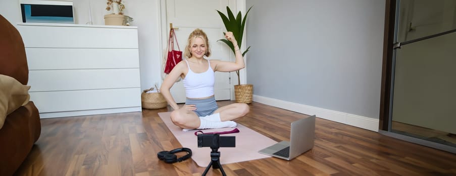 Portrait of young fitness instructor, vlogger showing exercises on camera, recording herself, sitting on mat with laptop, doing workout, explaining yoga movements to followers.