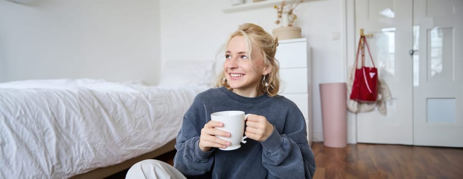 Lifestyle portrait of young woman sitting on bedroom floor with cup of tea, drinking from big white mug and looking aside, smiling happily.