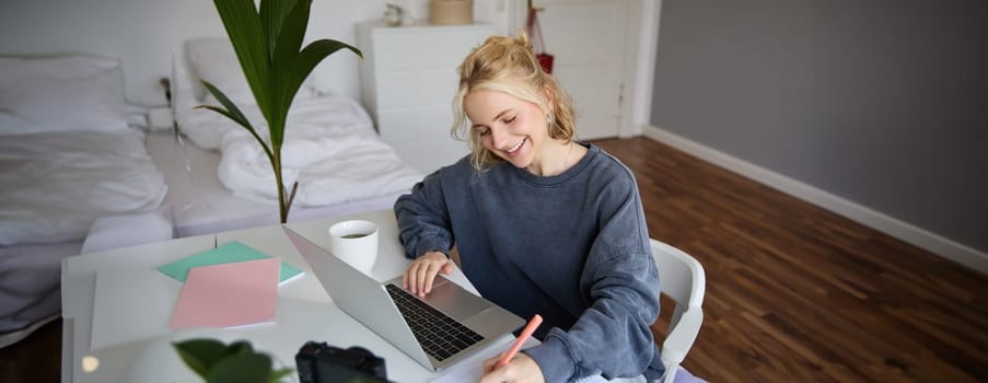 Portrait of smiling, beautiful blond woman, writing down notes, doing homework, studying from home, doing distance learning, online course, working remotely in her room.