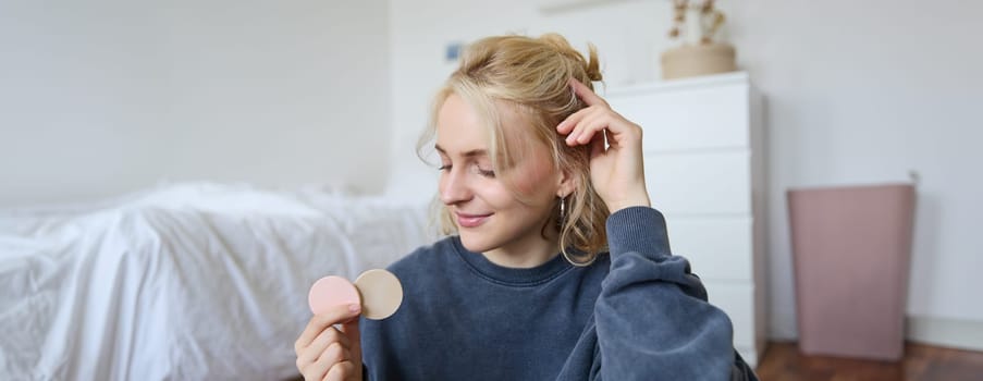 Portrait of young woman chatting on live stream about makeup, sits on floor in bedroom, showing beauty products to followers.