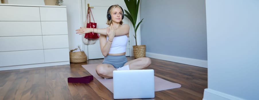 Wellbeing and fitness concept. Woman in headphones, sits on yoga mat, watches workout video on laptop, stretches arms, does exercises.