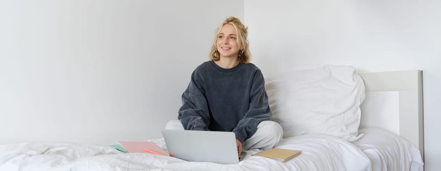 Portrait of young smiling woman studying in her bed, working from home in bedroom, sitting with laptop and notebooks on lotus pose, looking happy and relaxed.