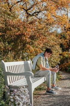 a teenager sits on a bench in the park drinks coffee from a thermo mug and looks into a phone. Portrait of handsome cheerful guy sitting on bench fresh air using device browsing media smm drinking latte urban outside outdoor.