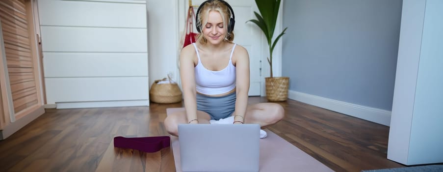 Portrait of young sporty woman following online video instructions during fitness workout, using laptop, sitting on yoga mat.