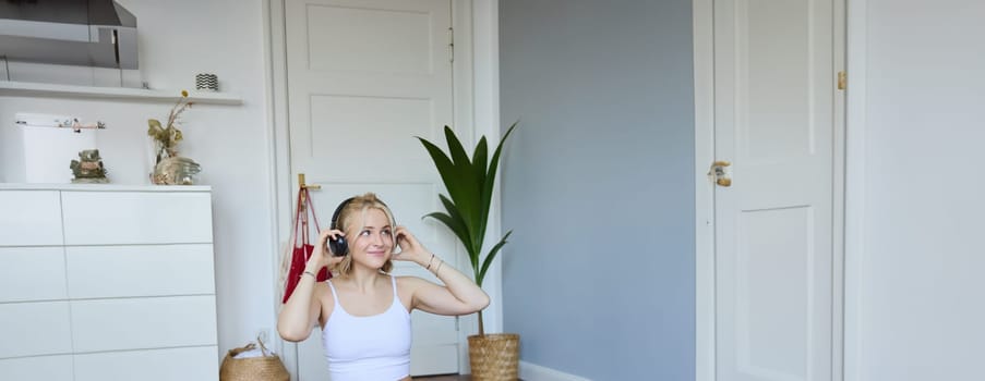 Portrait of cute young fitness woman, connects to online training session via laptop, wearing wireless headphones during workout, sits at home on rubber yoga mat.
