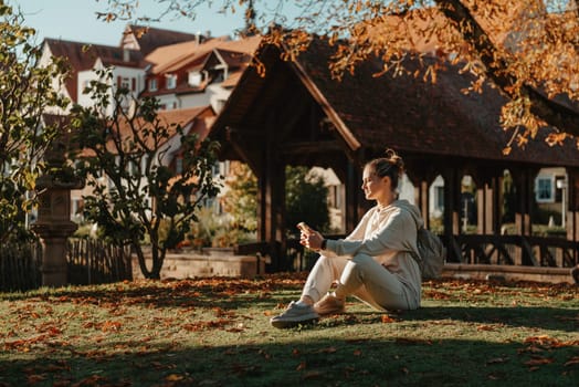 Young fashionable teenage girl with smartphone in park in autumn sitting at smiling. Trendy young woman in fall in park texting. Retouched, vibrant colors. Beautiful blonde teenage girl wearing casual modern autumn outfit sitting in park in autumn. Retouched, vibrant colors, brownish tones.