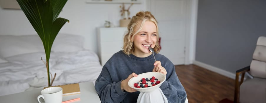 Close up portrait of smiling, cute blond woman, eating healthy lunch in her room, holding bowl with berries, biting spoon, looking happy.