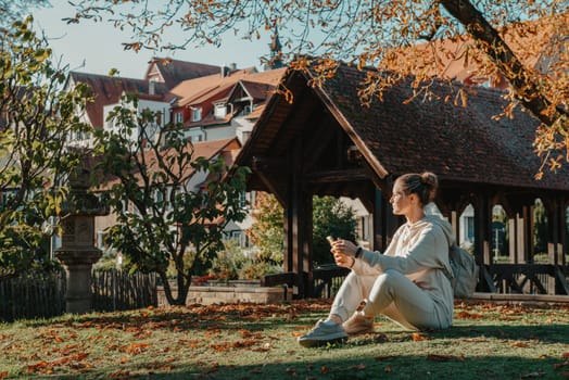 Young fashionable teenage girl with smartphone in park in autumn sitting at smiling. Trendy young woman in fall in park texting. Retouched, vibrant colors. Beautiful blonde teenage girl wearing casual modern autumn outfit sitting in park in autumn. Retouched, vibrant colors, brownish tones.