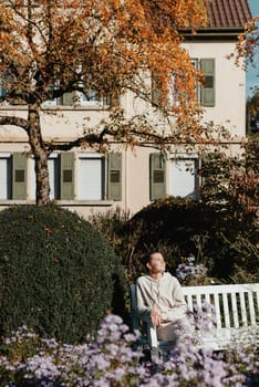 a girl sits on a bench in the park and enjoys the sun. Portrait young adult attractive woman enjoy sitting on bench and relaxing calm carefree rest in city park against green grass and trees on sunny day. Single female person relaxing chilling outdoors