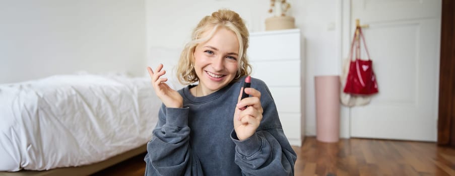 Portrait of cute, charismatic beauty blogger, woman sits in a room with lipstick in hand, talking about makeup, chatting with followers, recording online stream on social media app.