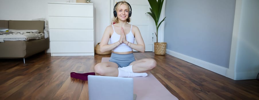 Portrait of young, relaxed woman in headphones, listens to meditation music on laptop, practice yoga on rubber mat, holding hands clasped together in namaste sign.