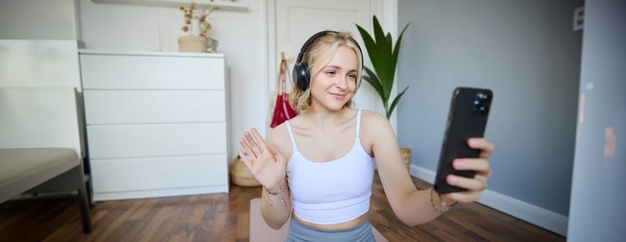 Portrait of young woman, fitness instructor doing online workout session on smartphone, waving hand at camera and saying hello, sitting on rubber mat in a room.