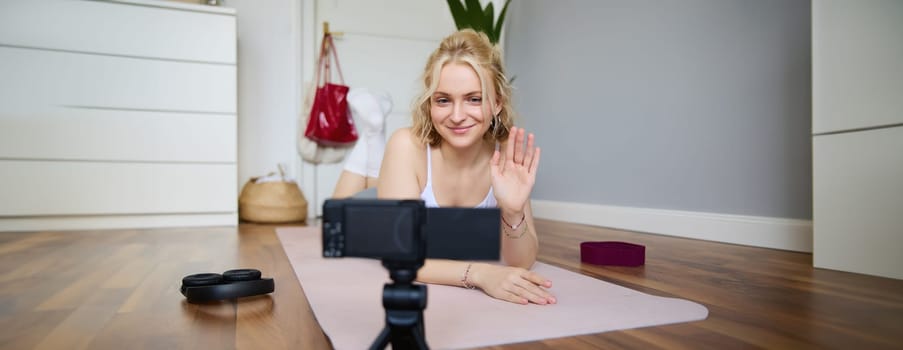 Portrait of smiling blond woman saying hello to her followers, fitness instructor recording video of herself showing workout exercises at home, lying on rubber yoga mat, looking at digital camera.