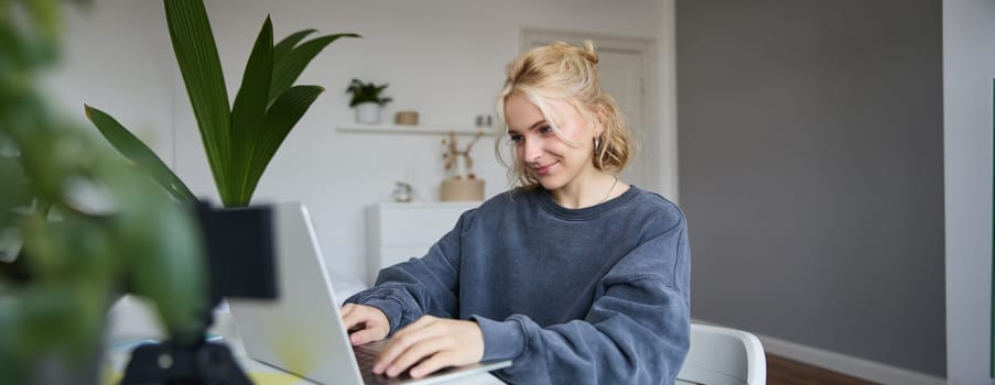 Portrait of young blond woman, female college student works from home on assignment, uses laptop, studies remotely, sits in room in front of computer.