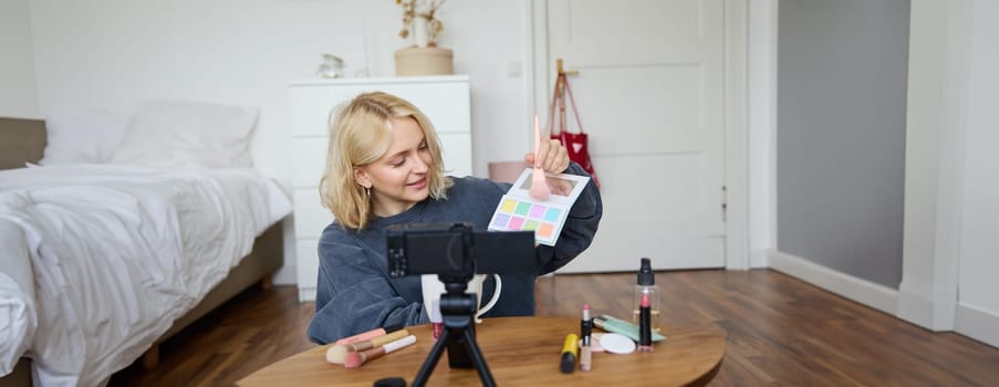 Portrait of smiling blond woman, teenage girl records a vlog on video camera, vlogger shows eyeshadows, tutorial how to put makeup on, sits in her room on floor near coffee table.