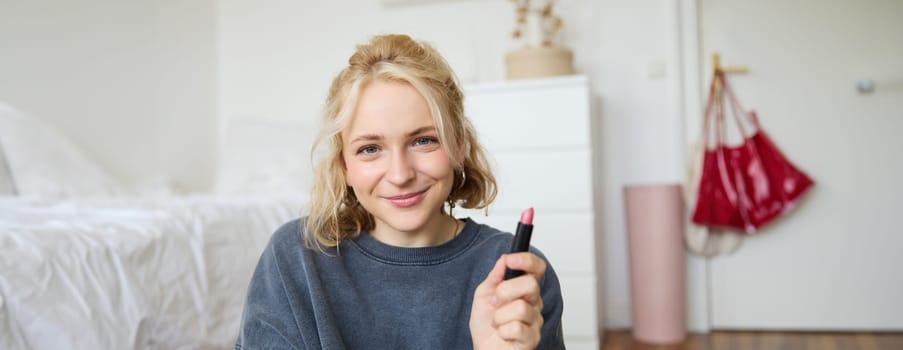 Portrait of smiling beautiful woman in her room, sitting and showing lipstick, recommending favourite beauty product, content maker recording a video of herself for social media blog.