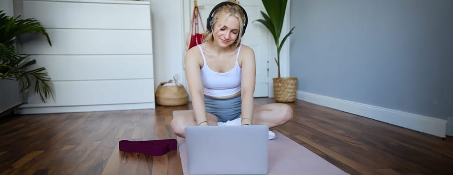 Portrait of smiling sporty woman at home, doing workout on rubber yoga mat, using laptop and wireless headphones.