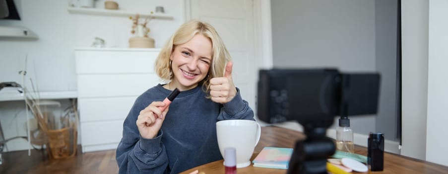 Portrait of smiling, beautiful young woman, lifestyle blogger recording a video in her room, sitting in front of camera on stabiliser, showing thumbs up and wink face, recommending mascara.