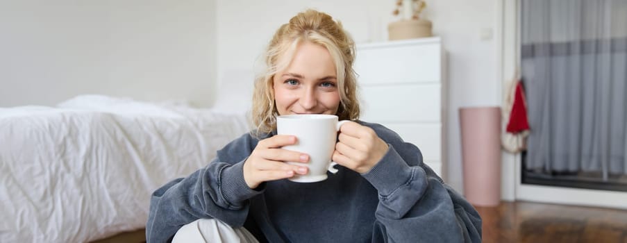 Image of young teenage girl sitting in her bedroom on floor, drinking cup of tea and enjoying day at home, smiling and looking at camera.