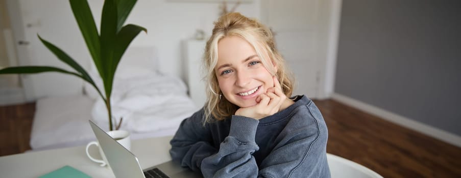 Close up portrait of young smiling female model, sits in a room with laptop, works from home, does online course, e-learning.