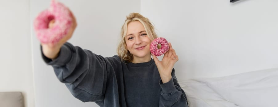 Portrait of beautiful smiling blond woman, showing two pink doughnuts at camera, eating delicious food.